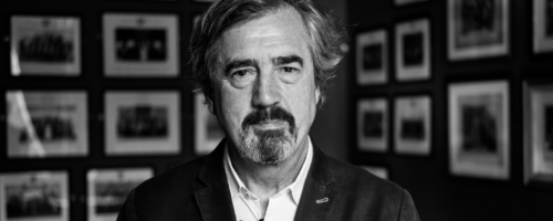 Irlandzki pisarz Sebastian Barry. Czarno-białe zdjęcie mężczyzny w średnim wieku z siwiejącymi włosami i brodą, ubranego w marynarkę i koszulę, stojącego na tle ściany z licznymi oprawionymi fotografiami (fot. Chris Boland / www.chrisboland.com CC BY-NC-ND 2.0)