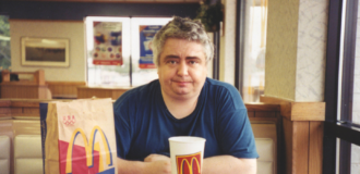 Fotografia. Mężczyzna (Daniel Johnston) siedzący przy stoliku w restauracji typu fast food, patrzy przed siebie. Ma siwe włosy i granatową koszulkę. Na stole przed nim znajduje się zestaw jedzenia z McDonald’s: tacka z frytkami i pudełkiem burgera, duży kubek z napojem oraz papierowa torba z widocznym logo „M”. Wnętrze lokalu ma jasne oświetlenie, tapicerowane siedzenia i plakaty na ścianach w tle.