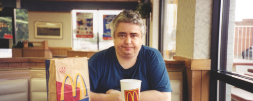 Fotografia. Mężczyzna (Daniel Johnston) siedzący przy stoliku w restauracji typu fast food, patrzy przed siebie. Ma siwe włosy i granatową koszulkę. Na stole przed nim znajduje się zestaw jedzenia z McDonald’s: tacka z frytkami i pudełkiem burgera, duży kubek z napojem oraz papierowa torba z widocznym logo „M”. Wnętrze lokalu ma jasne oświetlenie, tapicerowane siedzenia i plakaty na ścianach w tle.