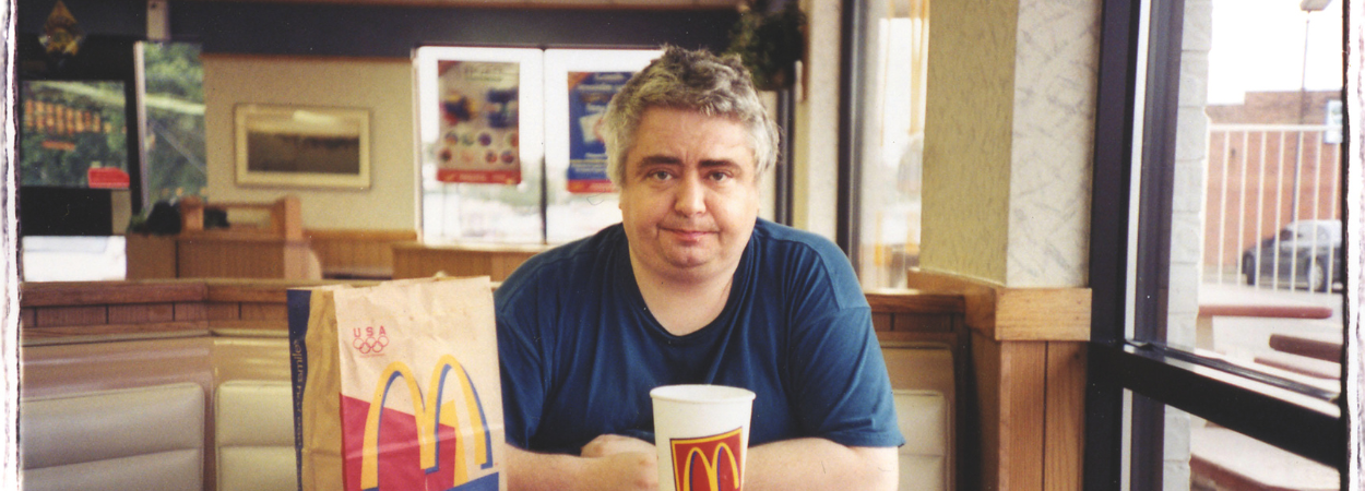 Fotografia. Mężczyzna (Daniel Johnston) siedzący przy stoliku w restauracji typu fast food, patrzy przed siebie. Ma siwe włosy i granatową koszulkę. Na stole przed nim znajduje się zestaw jedzenia z McDonald’s: tacka z frytkami i pudełkiem burgera, duży kubek z napojem oraz papierowa torba z widocznym logo „M”. Wnętrze lokalu ma jasne oświetlenie, tapicerowane siedzenia i plakaty na ścianach w tle.