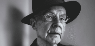 Czarno-biała fotografia. Starszy mężczyzna (Jacek Kubitsky) na szarym tle. Ma cienkie okulary, patrzy przed siebie. Jest ubrany w czarny kapelusz oaz czarną marynarkę. W uniesionej dłoni trzyma papierosa