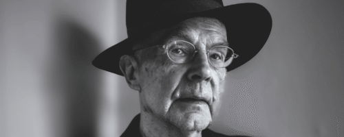 Czarno-biała fotografia. Starszy mężczyzna (Jacek Kubitsky) na szarym tle. Ma cienkie okulary, patrzy przed siebie. Jest ubrany w czarny kapelusz oaz czarną marynarkę. W uniesionej dłoni trzyma papierosa