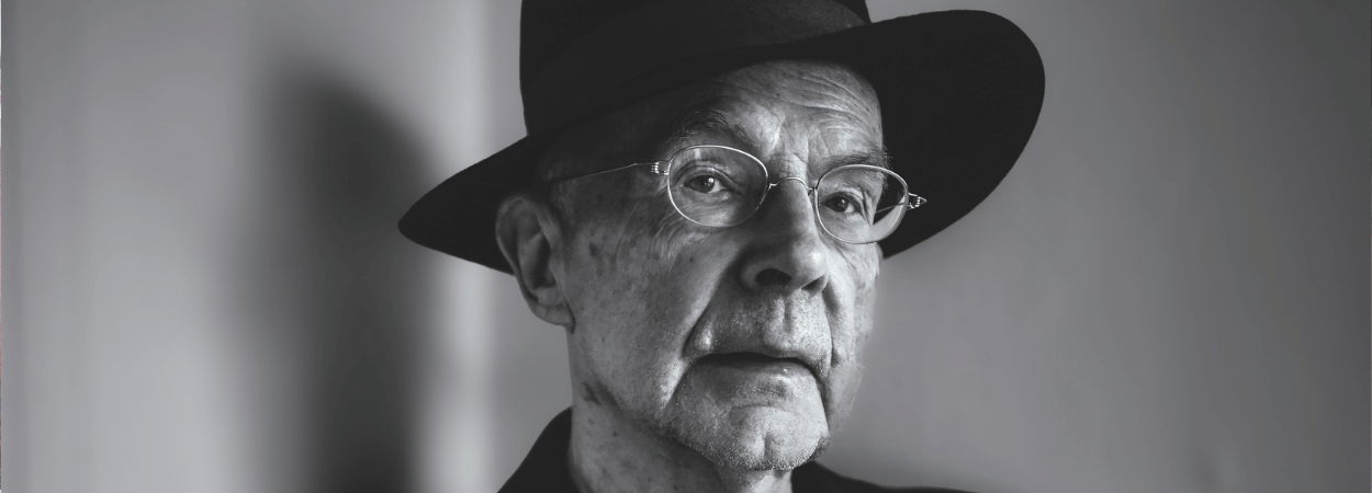 Czarno-biała fotografia. Starszy mężczyzna (Jacek Kubitsky) na szarym tle. Ma cienkie okulary, patrzy przed siebie. Jest ubrany w czarny kapelusz oaz czarną marynarkę. W uniesionej dłoni trzyma papierosa