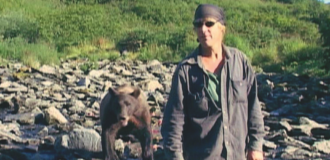 Kadr z filmu "Grizzly Man". Górski krajobraz. Na pierwszym planie znajduje się mężczyzna w zabrudzonej pyłem, ciemnej koszuli. Na głowie ma chustkę. Nosi ciemnie okulary. Za nim stoi brunatny niedźwiedź. Oboje są w kamienistym strumyku.