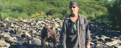 Kadr z filmu "Grizzly Man". Górski krajobraz. Na pierwszym planie znajduje się mężczyzna w zabrudzonej pyłem, ciemnej koszuli. Na głowie ma chustkę. Nosi ciemnie okulary. Za nim stoi brunatny niedźwiedź. Oboje są w kamienistym strumyku.
