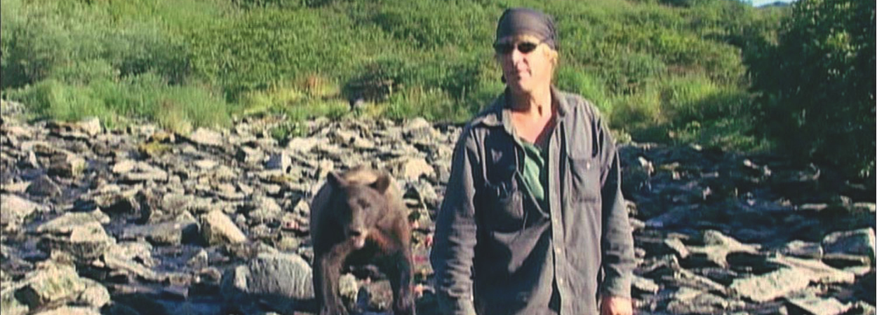 Kadr z filmu "Grizzly Man". Górski krajobraz. Na pierwszym planie znajduje się mężczyzna w zabrudzonej pyłem, ciemnej koszuli. Na głowie ma chustkę. Nosi ciemnie okulary. Za nim stoi brunatny niedźwiedź. Oboje są w kamienistym strumyku.