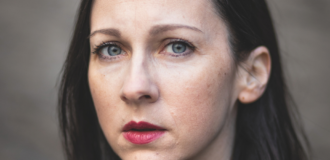 Fotografia. Portret kobiety (Kristina Kocan) na jednolitym, rozmytym tle. Ma włosy do ramion, i patrzy bezpośrednio w obiektyw. Ubrana jest w ciemny płaszcz lub marynarkę z dużym kołnierzem.