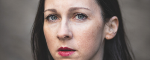 Fotografia. Portret kobiety (Kristina Kocan) na jednolitym, rozmytym tle. Ma włosy do ramion, i patrzy bezpośrednio w obiektyw. Ubrana jest w ciemny płaszcz lub marynarkę z dużym kołnierzem.